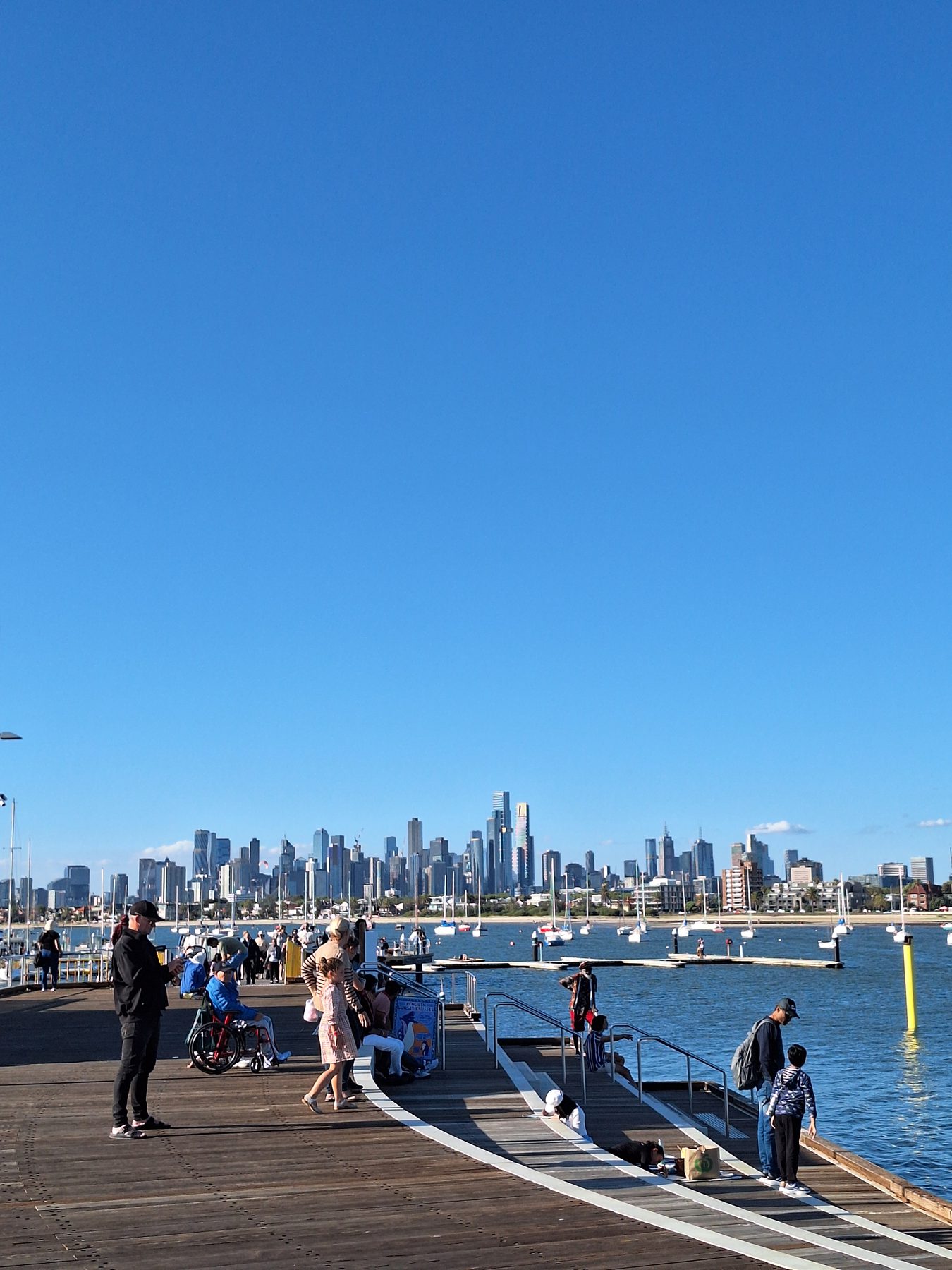 The Melbourne city skyline seen across the water from St Kilda pier, with people walking along the boardwalk