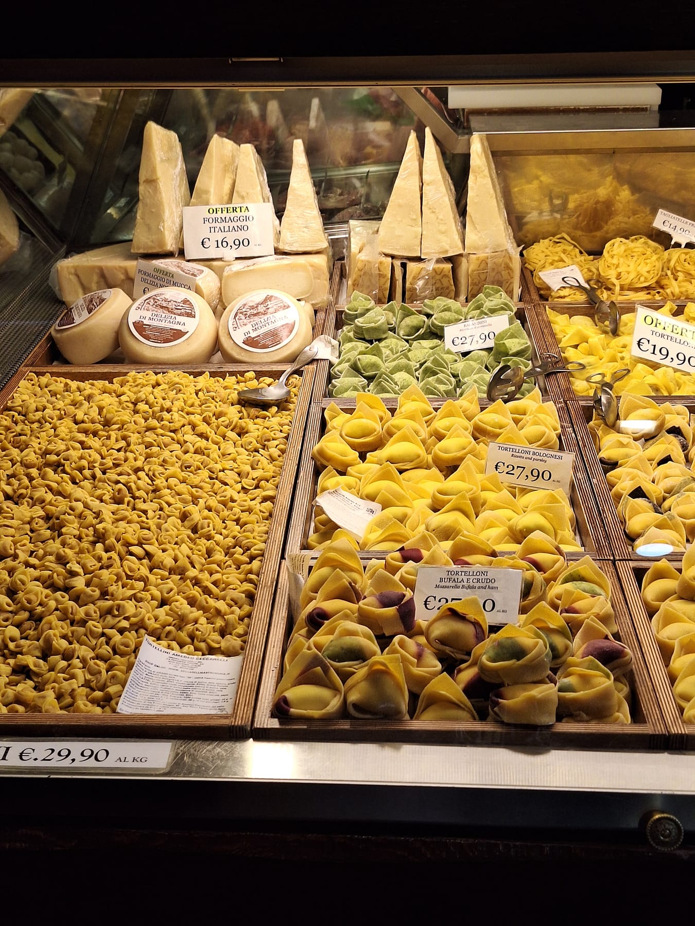 A shop counter in Bologna piled with trays of fresh tortellini and tortelloni, with wedges of cheese behind