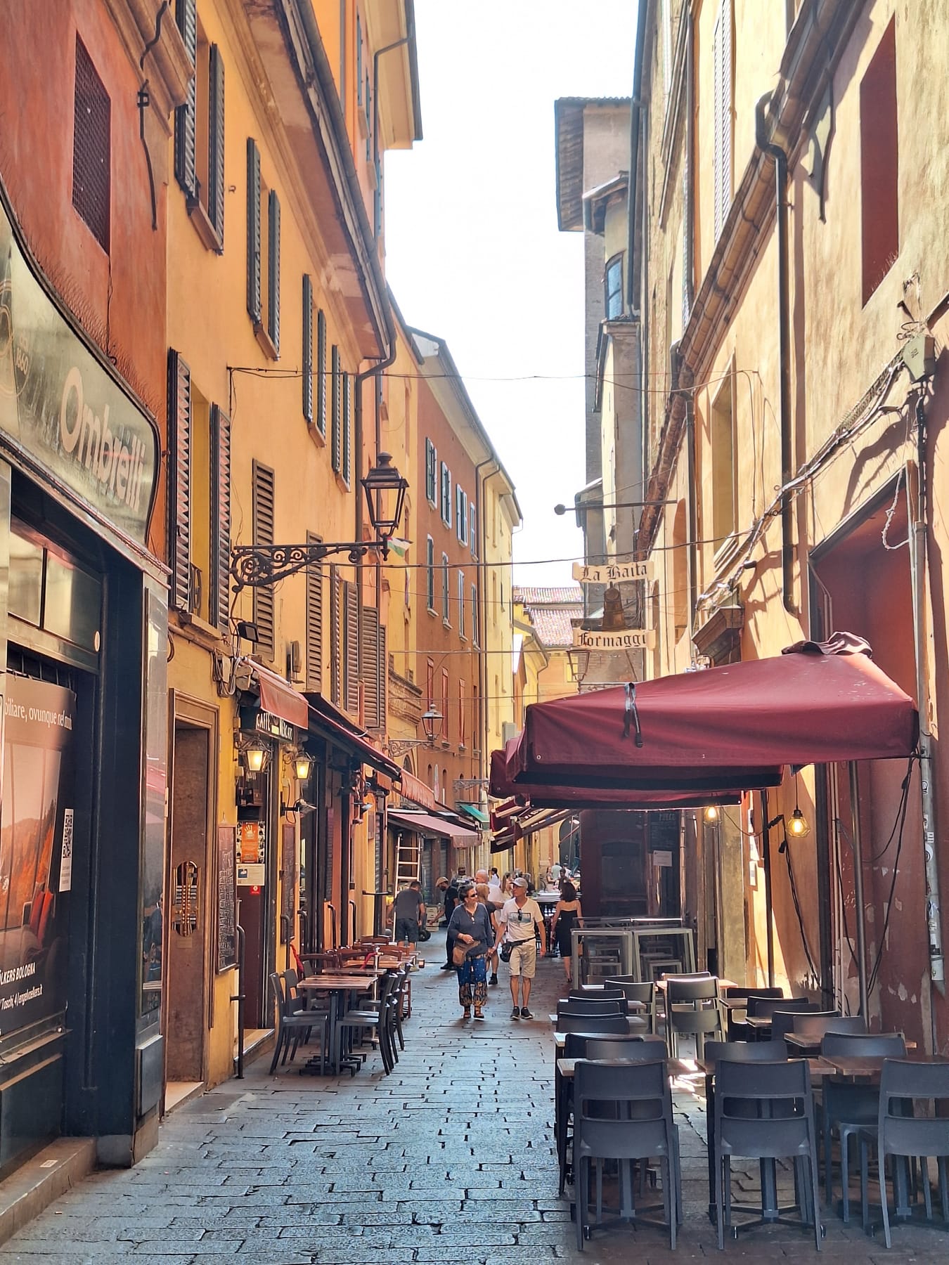 A narrow cobbled lane in the Quadrilatero with café tables, red awnings and a hanging sign for a cheese shop