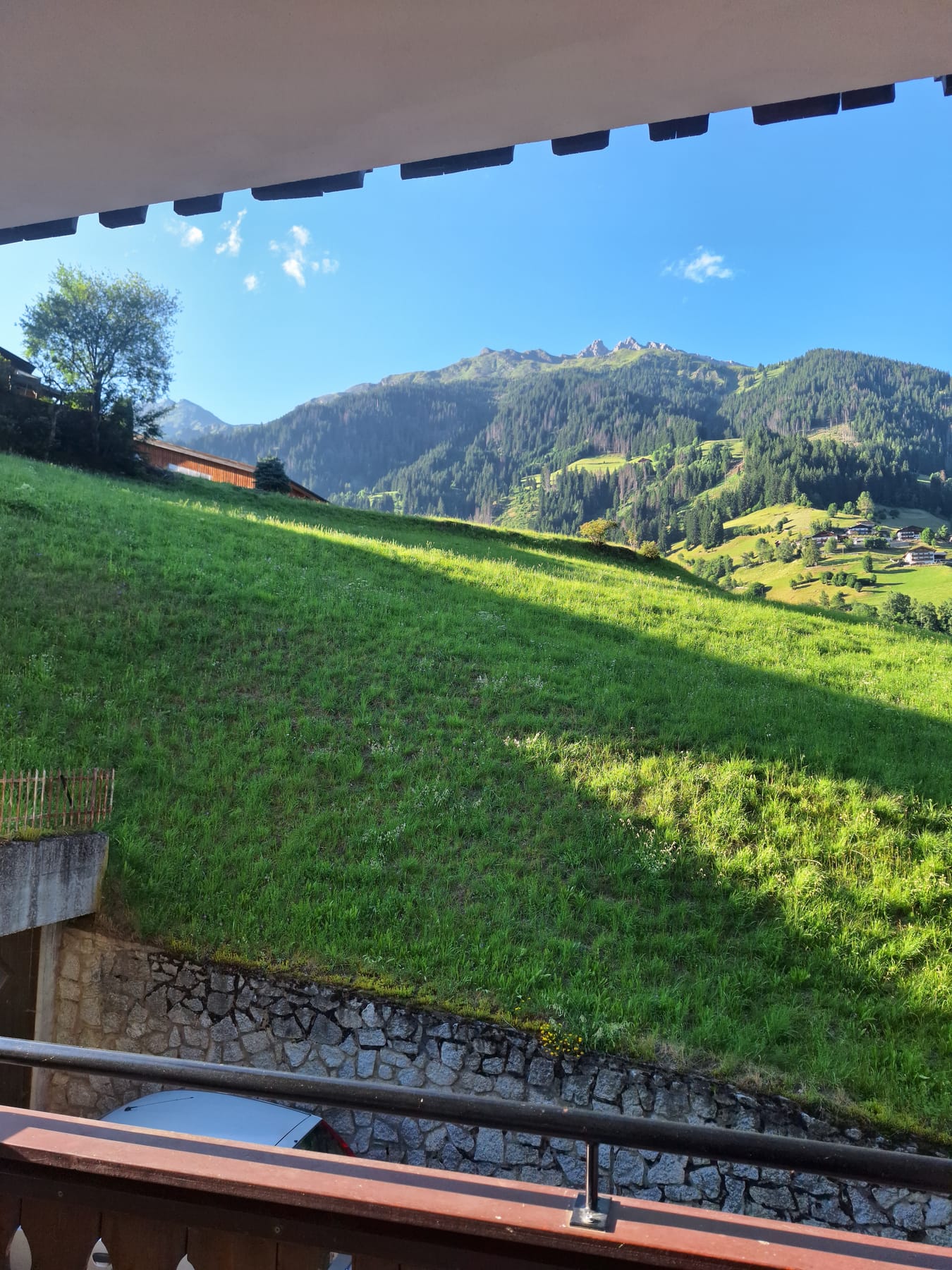 A view from a wooden balcony over steep green pasture and forested mountains in evening sun