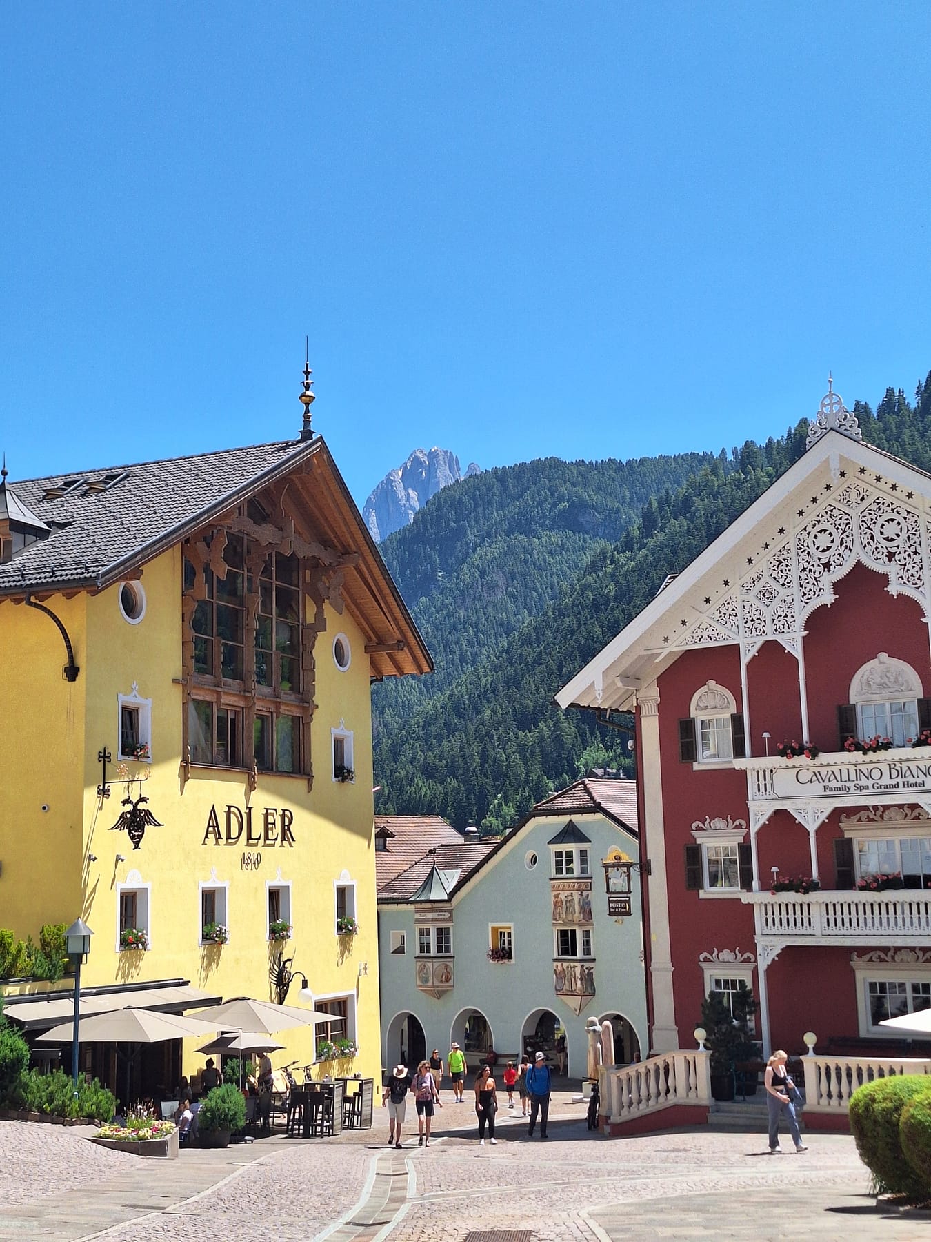 Painted Alpine hotels in Ortisei, one yellow and one red with white carved balconies, under a rocky peak