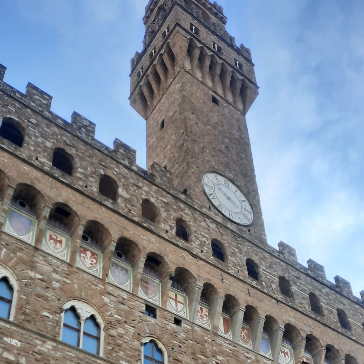 The crenellated clock tower and stone façade of the Palazzo Vecchio in Florence against a blue sky