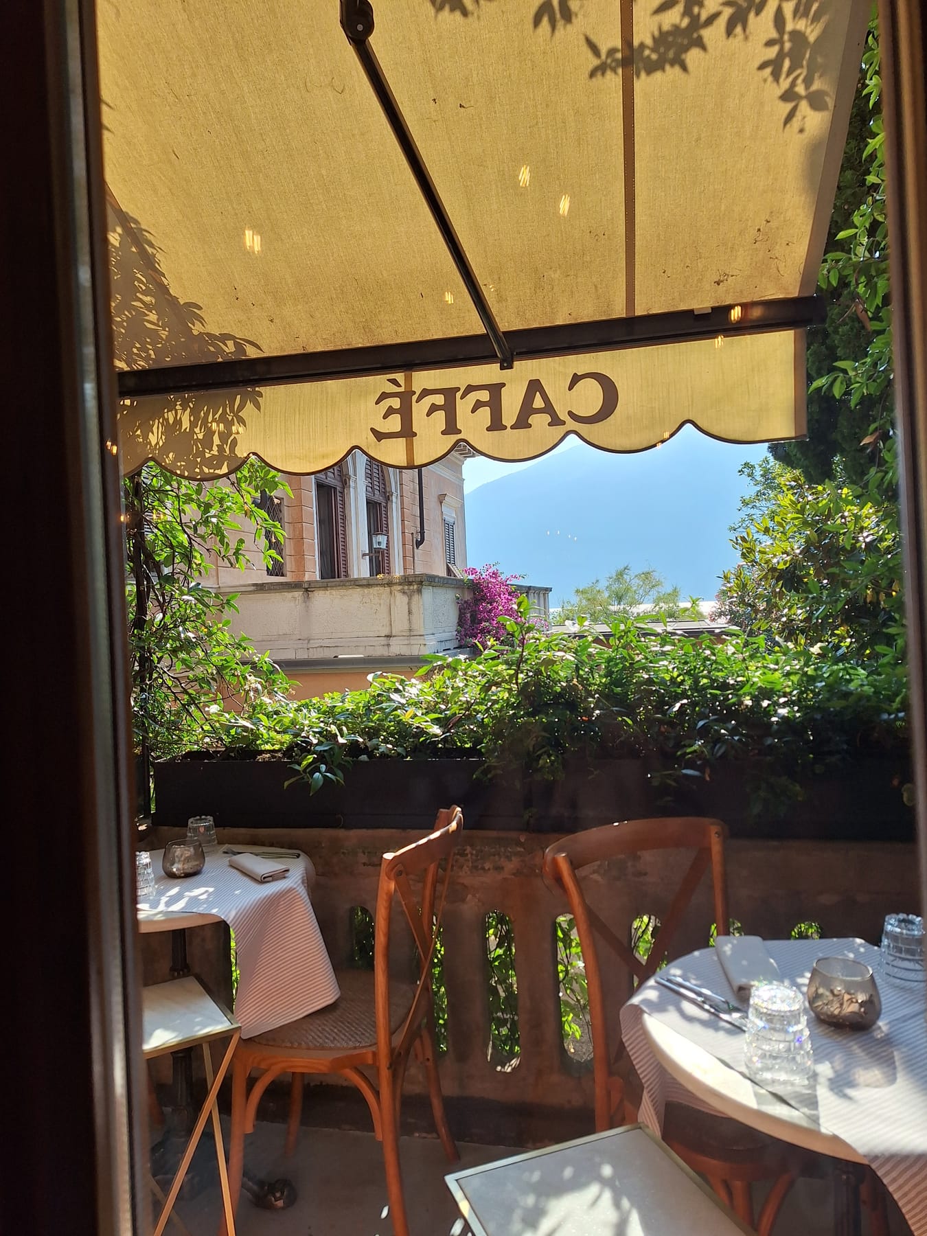 A café terrace with wooden chairs and white tables under an awning, looking out through greenery to the lake