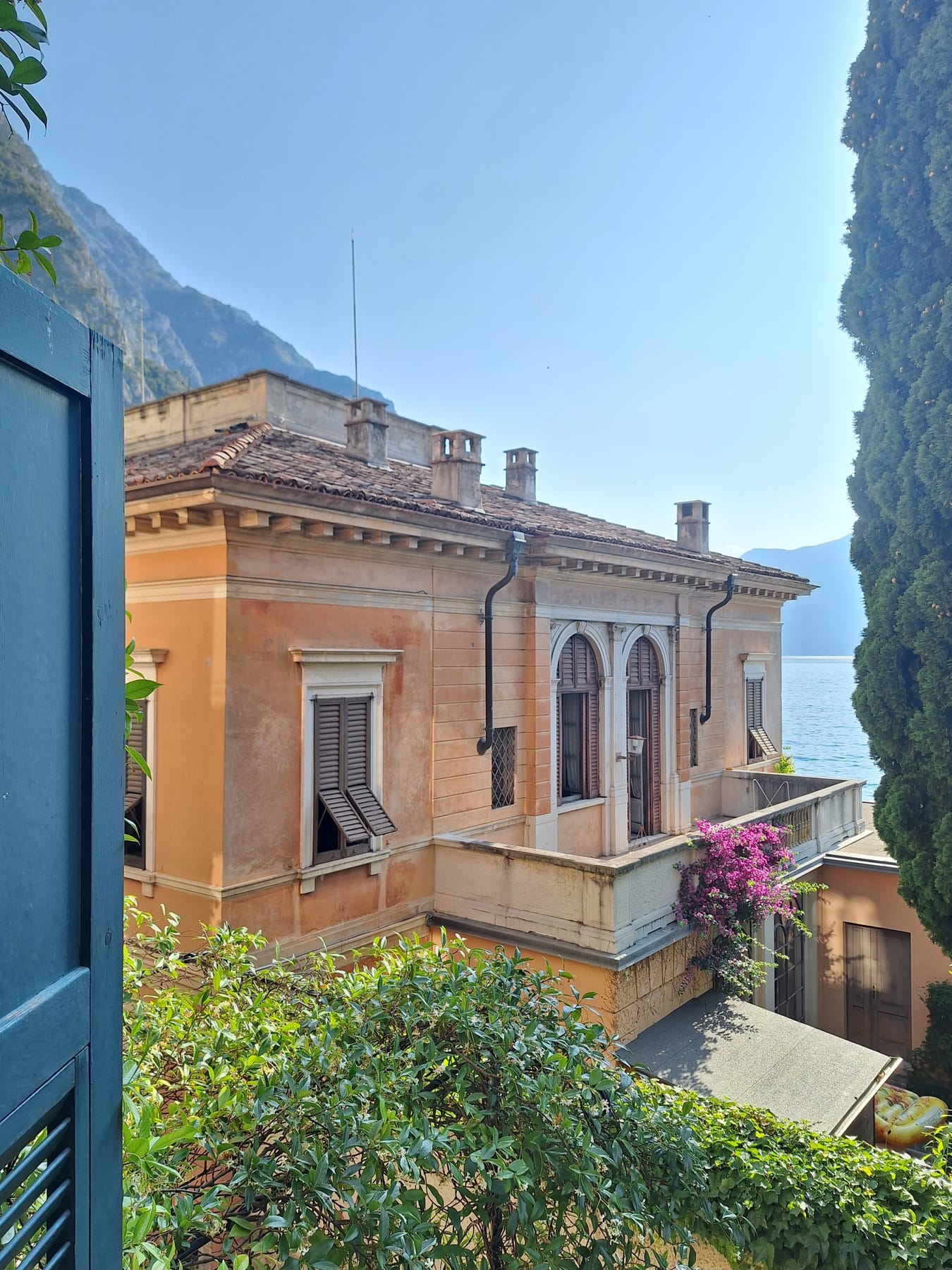 An ochre villa with arched windows and bougainvillea on its balcony, with Lake Garda and mountains behind