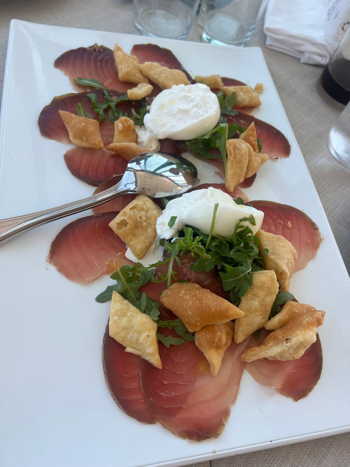 Thin slices of tuna carpaccio topped with burrata, rocket and crisp fried pasta on a white plate