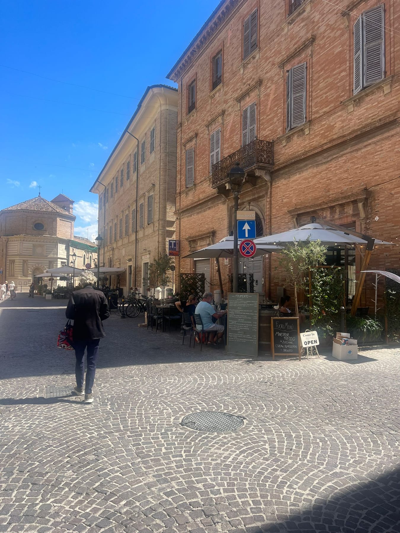 A cobbled street of brick palazzi with café umbrellas and a church dome at the end, under a blue sky