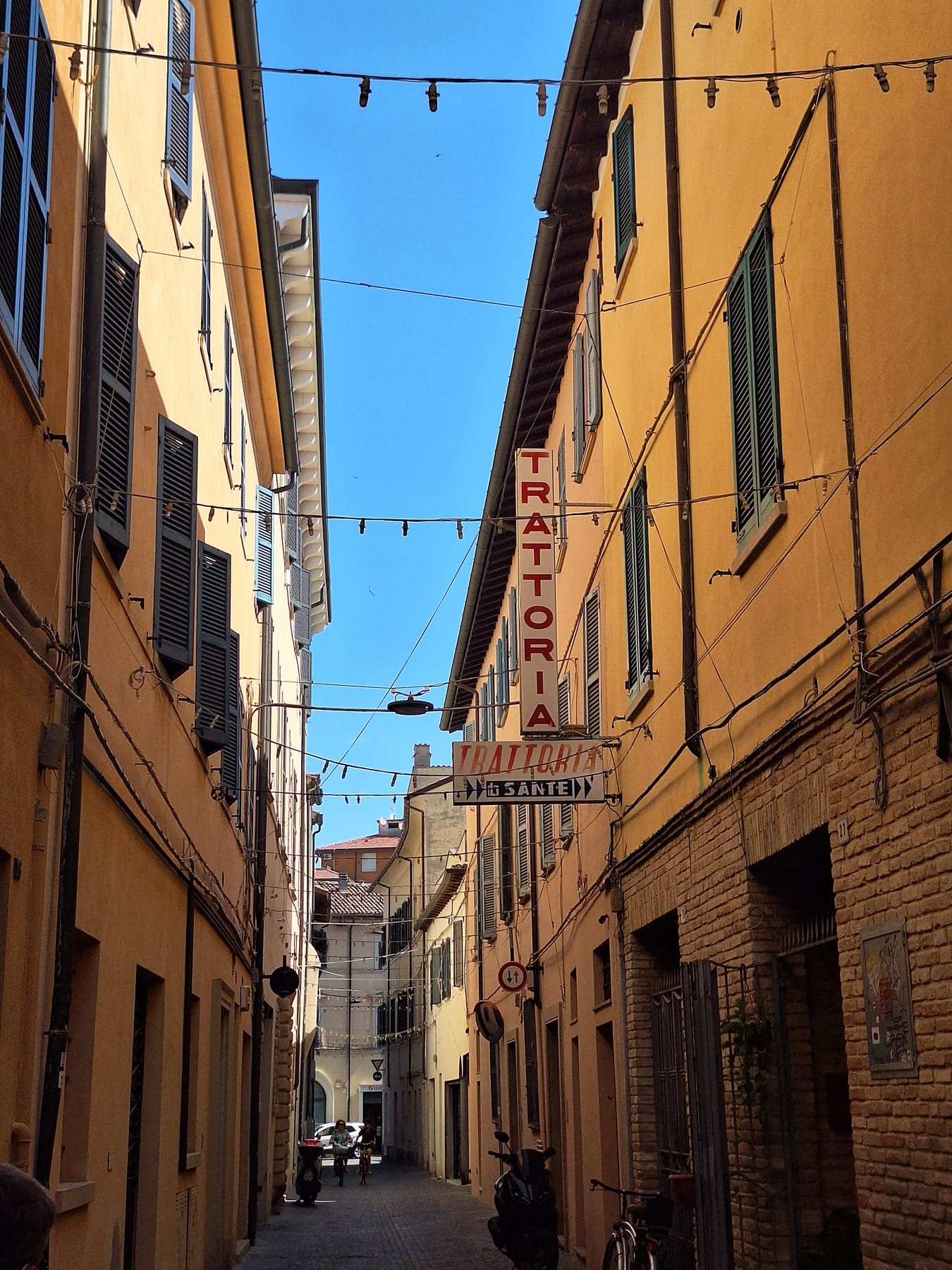 A narrow street of ochre buildings with a vertical trattoria sign and bunting strung overhead