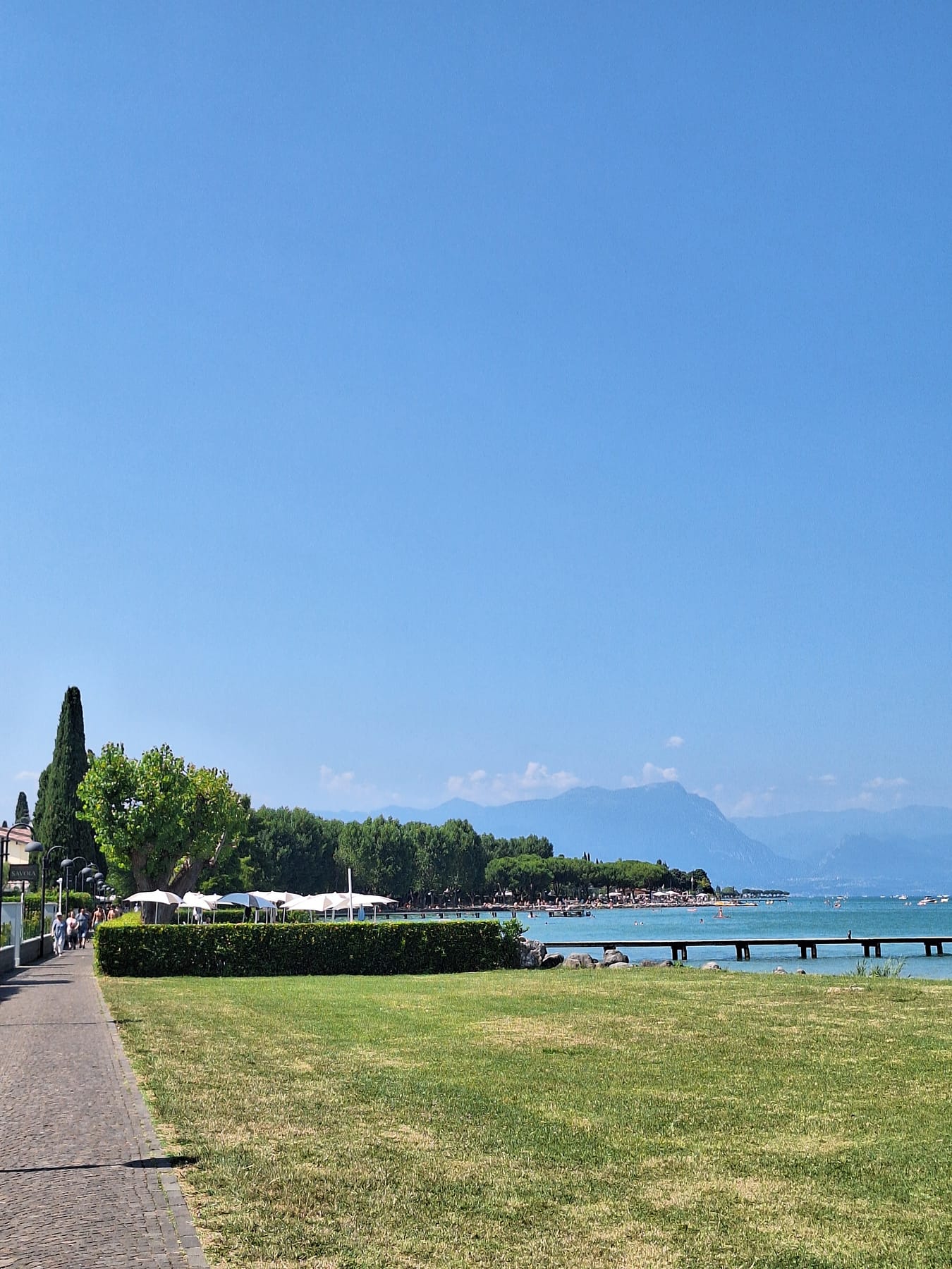 A lakeside lawn and path lined with trees, with a jetty, turquoise water and mountains across Lake Garda