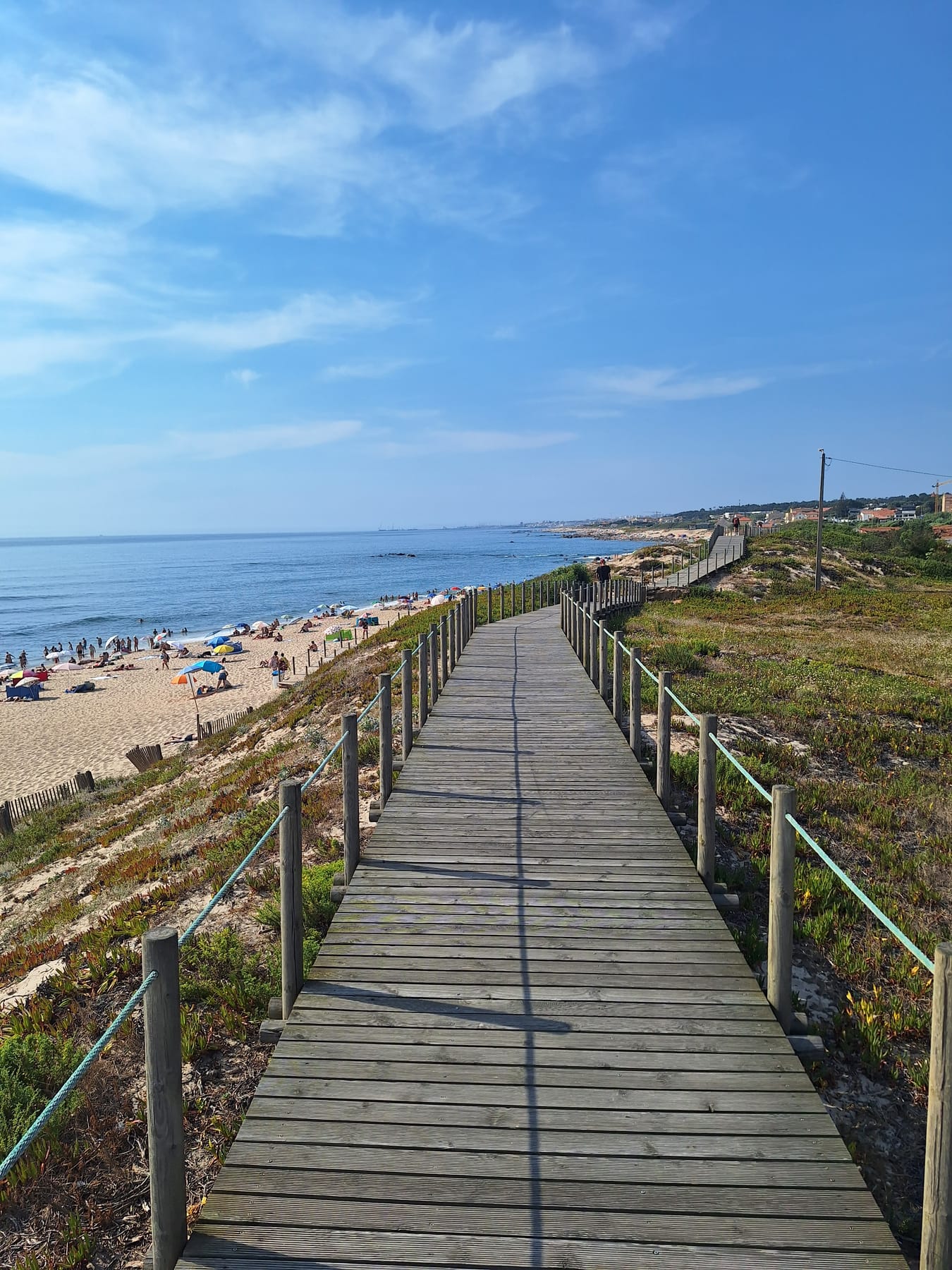 A wooden boardwalk running through grassy dunes beside a busy sandy beach on the Atlantic coast near Porto