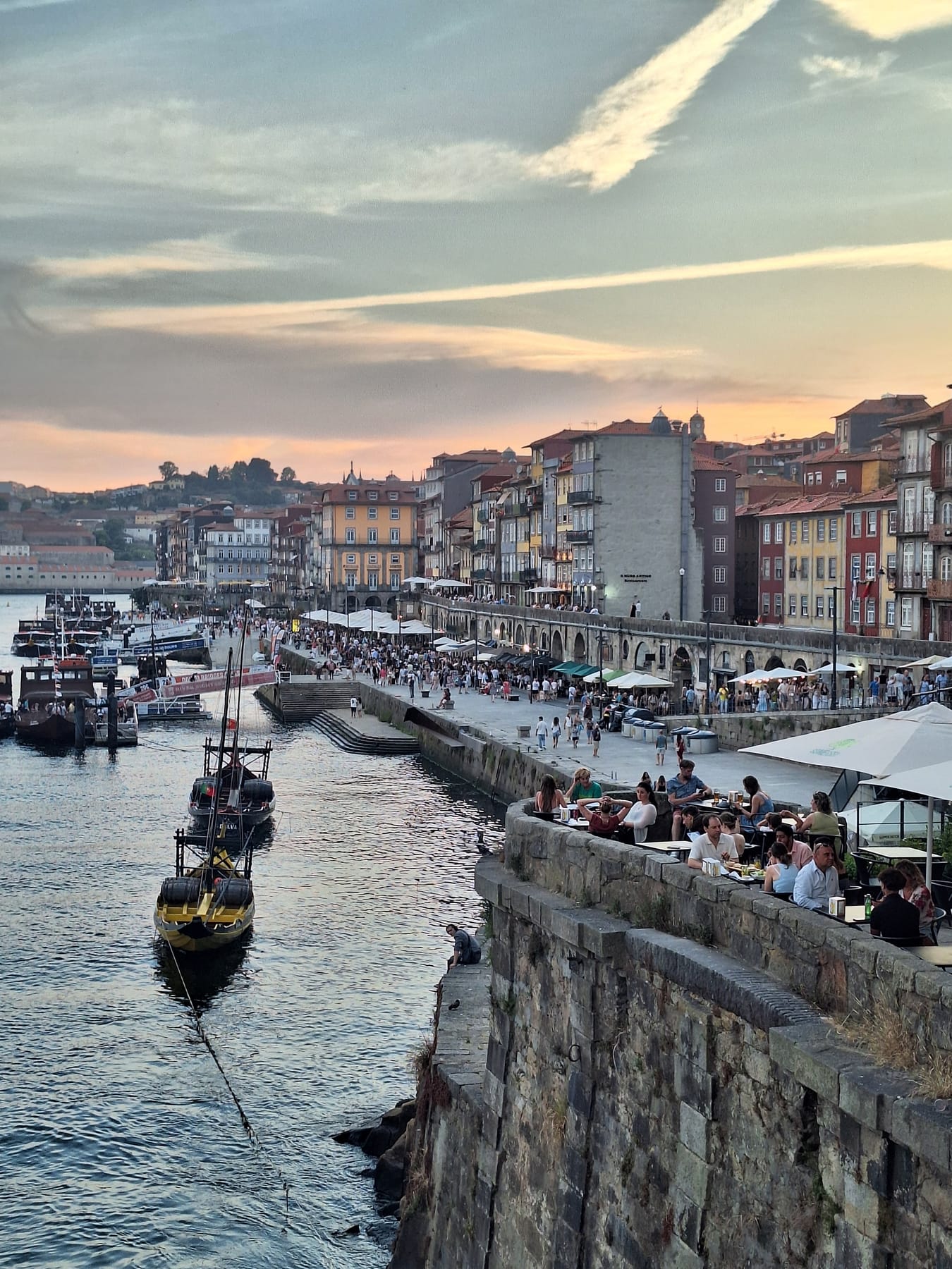 Crowded riverside terraces along the Ribeira in Porto at sunset, with traditional boats moored on the Douro