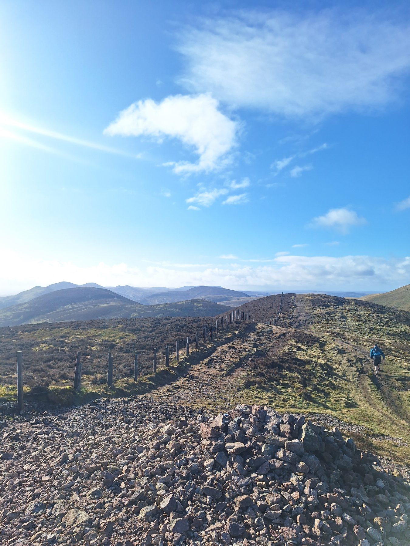 A rocky hilltop path with hills stretching into the distance under a blue sky near Edinburgh